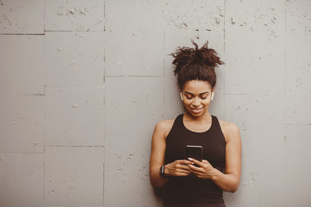Happy african american girl in sportswear with smartwatch and wireless headphones looks at smartphoneの写真素材