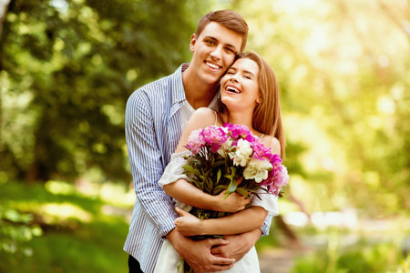 First anniversary gift. Millennial married couple with bouquet of flowers hugging at parkの写真素材