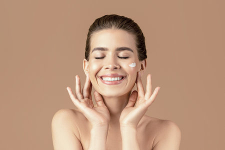 Lovely young lady with perfect skin touching her face, applying moisturizing cream on brown studio backgroundの写真素材