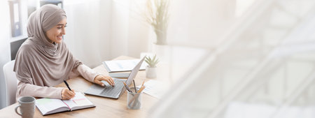 Muslim woman in hijab using laptop and taking notes in officeの写真素材