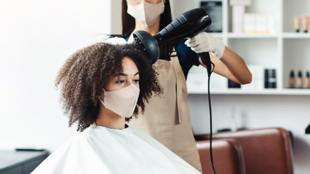 Young hairdresser in protective mask making stylish hairdo for customerの写真素材