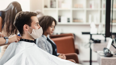 Young man in protective mask sitting in client chair at barber shop, waiting for haircut