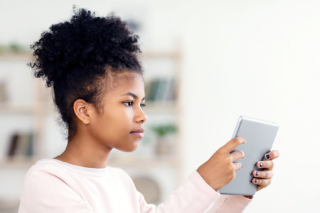 African Preteen Girl Using Digital Tablet Reading Book At Homeの写真素材