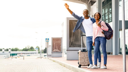 Happy Young Black Couple Standing With Suitcases Near Airport Terminal, Catching Taxiの写真素材