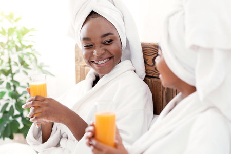 Two joyful black women drinking fresh juice in bedの写真素材