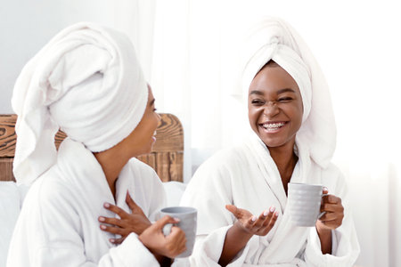 Happy girls in bathrobes sitting on bed and drinking coffeeの写真素材
