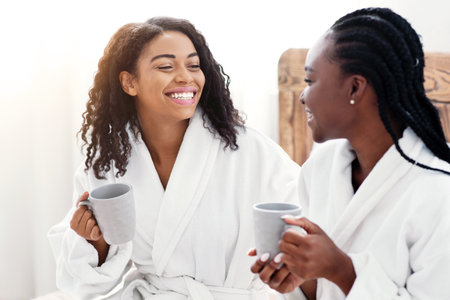 Happy girlfriends in bathrobes sitting on bed and drinking coffeeの写真素材