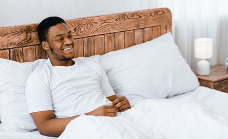 Happy Relaxed African American Man Sitting In Bed In Bedroomの写真素材