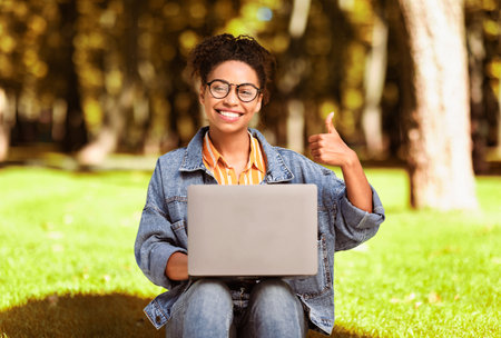African Woman Working On Laptop Gesturing Thumbs-Up Sitting In Parkの写真素材