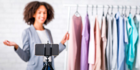 Modern technology for work online from home during self isolating. Blurred happy african american business woman presenting collection of clothes and recording video on smartphoneの写真素材