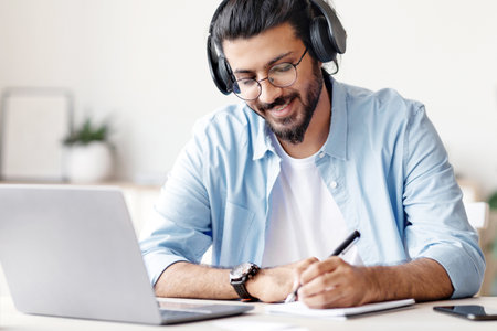 Eastern Guy Wearing Headphones Studying Online With Laptop At Home, Taking Notes