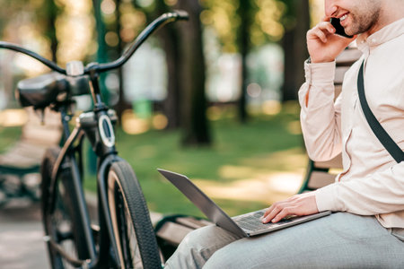 Young man enjoys working on laptop while talking on phone in the parkの写真素材