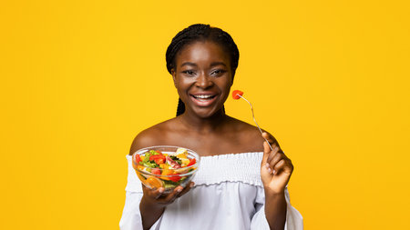 Dieting Concept. Joyful african american lady enjoying fresh vegetable salad, yellow backgroundの写真素材