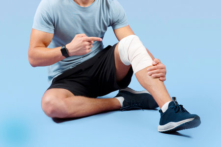 Athletic millennial man having sports injury, pointing at his sprained bandaged knee over lilac...