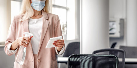 Return to work, workplace hygiene and new normal. Blond woman in business suit in protective mask with paper towel and spray disinfect table in modern officeの写真素材