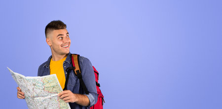 Cheerful male tourist with backpack holding map and looking aside at empty space over blue studio backgroundの写真素材