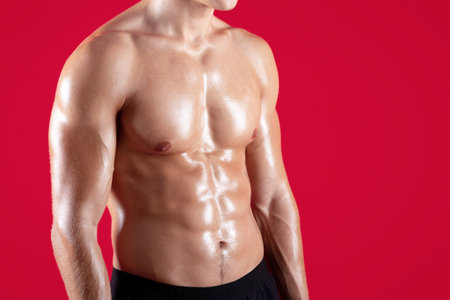 Closeup view of young strong sportsman with naked torso and prominent abs muscles on red studio...