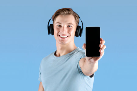 Positive young athlete in headphones showing cellphone with blank screen over violet background,...