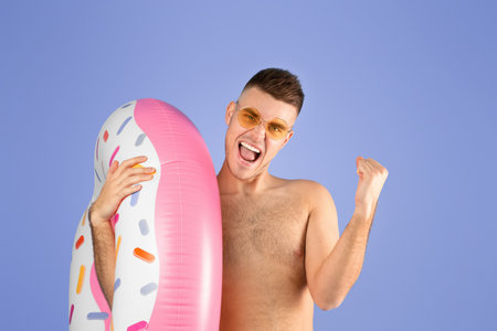 Thrilled young man in shorts holding inflatable ring and making YES gesture over blue studio backgroundの写真素材