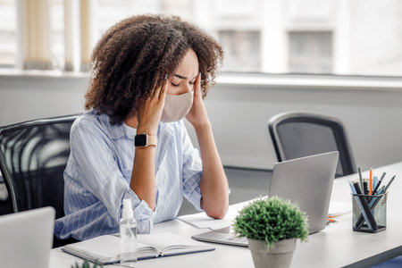 Overwork, headache and first signs of disease. African american woman in protective mask suffering from headache, sitting at table with laptop and antisepticの写真素材