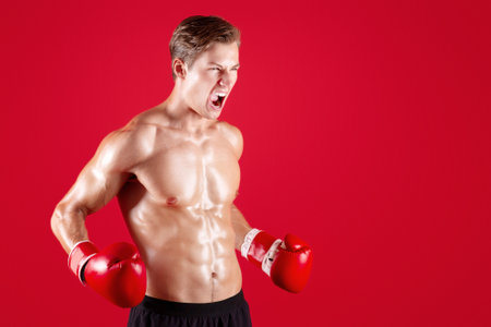 Aggressive young sportsman in boxing gloves screaming in anger on red studio backgroundの写真素材