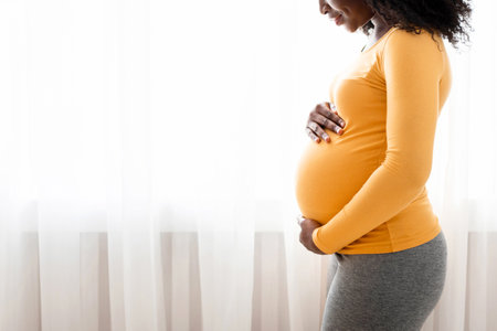 Beautiful pregnant black woman hugging her tummy at homeの写真素材