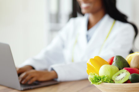 Nutritionist working on laptop, giving online consultation to client at clinic, focus on bowl of fruits and vegetablesの写真素材