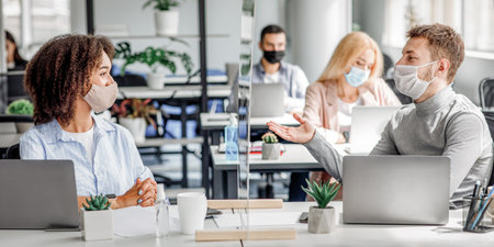 Communication and meeting in office after returning from quarantine. Young guy and african american woman in protective mask talking through glass board at workplaceの写真素材