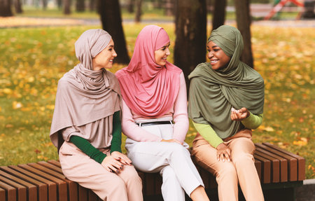 Three Muslim Ladies In Hijab Talking Sitting On Bench Outdoorsの写真素材