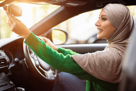 Happy muslim woman driving alone adjusting rearview mirrorの写真素材
