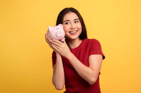 A young woman joyfully holds a pink piggy bank near her cheek, exuding happinessの写真素材