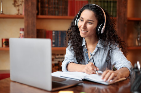 Woman sitting at desk, using laptop and writing in notebook