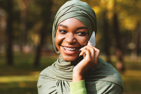 Cheerful African American Muslim Female Talking On Cellphone Standing Outdoors