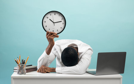 Tired african american businessman sitting at desk with pc laptop, holding clock, feeling exhausted,...