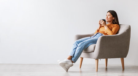 Relaxed millennial Asian woman drinking coffee, having break, chilling in armchair against white wall, free spaceの写真素材