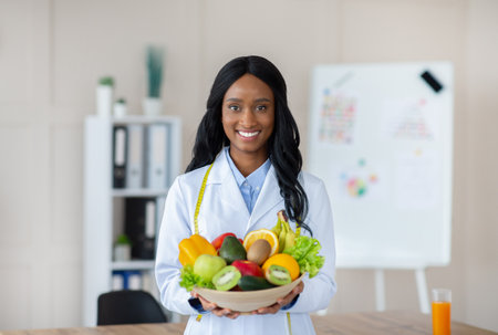 Portrait of happy black dietitian in lab coat holding bowl of fresh fruits and vegetables, smiling at camera at clinicの写真素材