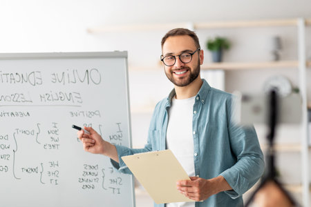 Portrait of happy young male teacher standing near blackboard with English grammar rules, conducting...