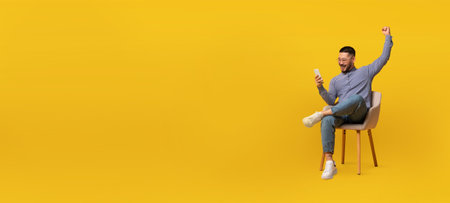 Man celebrates joyfully while using a smartphone against a yellow backgroundの写真素材