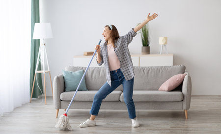Happy woman singing while washing floor at living room, using mop as microphone, having fun during...