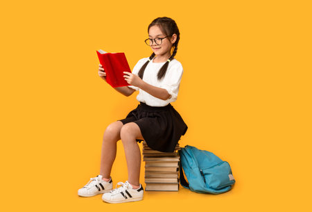 Cheerful Asian Schoolgirl Reading Sitting On Book Stack In Studioの写真素材