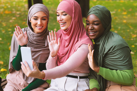 Muslim Ladies Making Video Call Using Phone Waving Hands Outdoorsの写真素材