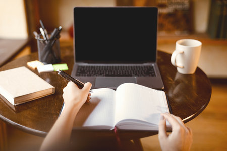 Woman sitting at desk using laptop, writing on notebook, povの写真素材
