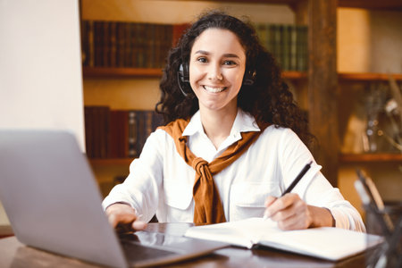 Woman sitting at desk, using laptop and writing in notebook