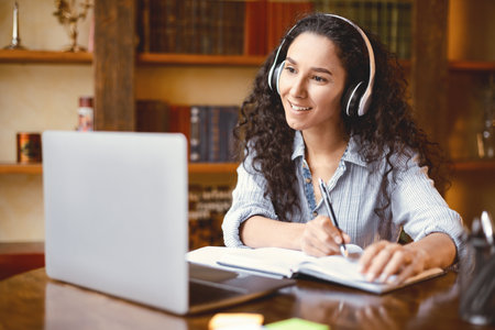 Woman sitting at desk, using laptop and writing in notebook