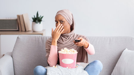 Young black woman in hijab watching scary movie, covering her mouth in horror, eating popcorn on sofa at homeの写真素材