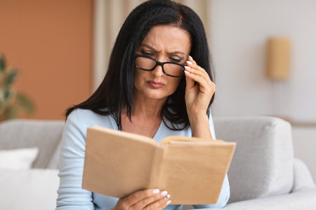 Mature woman touching glasses trying to read bookの写真素材