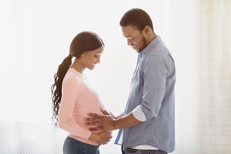 Side view of handsome black man touching his wifes pregnant belly near window at home