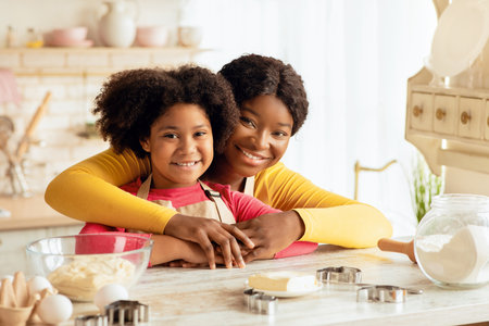 Loving Black Mom And Daughter In Aprons Posing At Table In Kitchenの写真素材