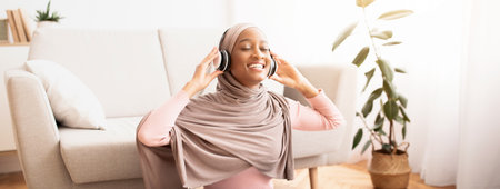 Full length portrait of black Muslim woman in hijab and headphones listening to music on floor in living roomの写真素材