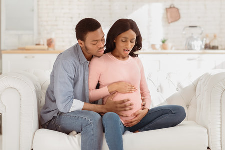 African American pregnant woman suffering from pain, having birth contractions while worried husband helping her at homeの写真素材
